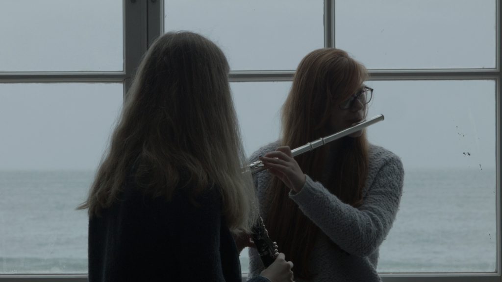 A film still showing two young women in front of a window, playing a flute and a clarinet